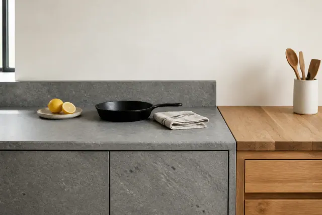 Daylit kitchen corner where a honed gray stone run meets a wood block section, with a cut lemon, a cast iron skillet and a folded towel on the counter
