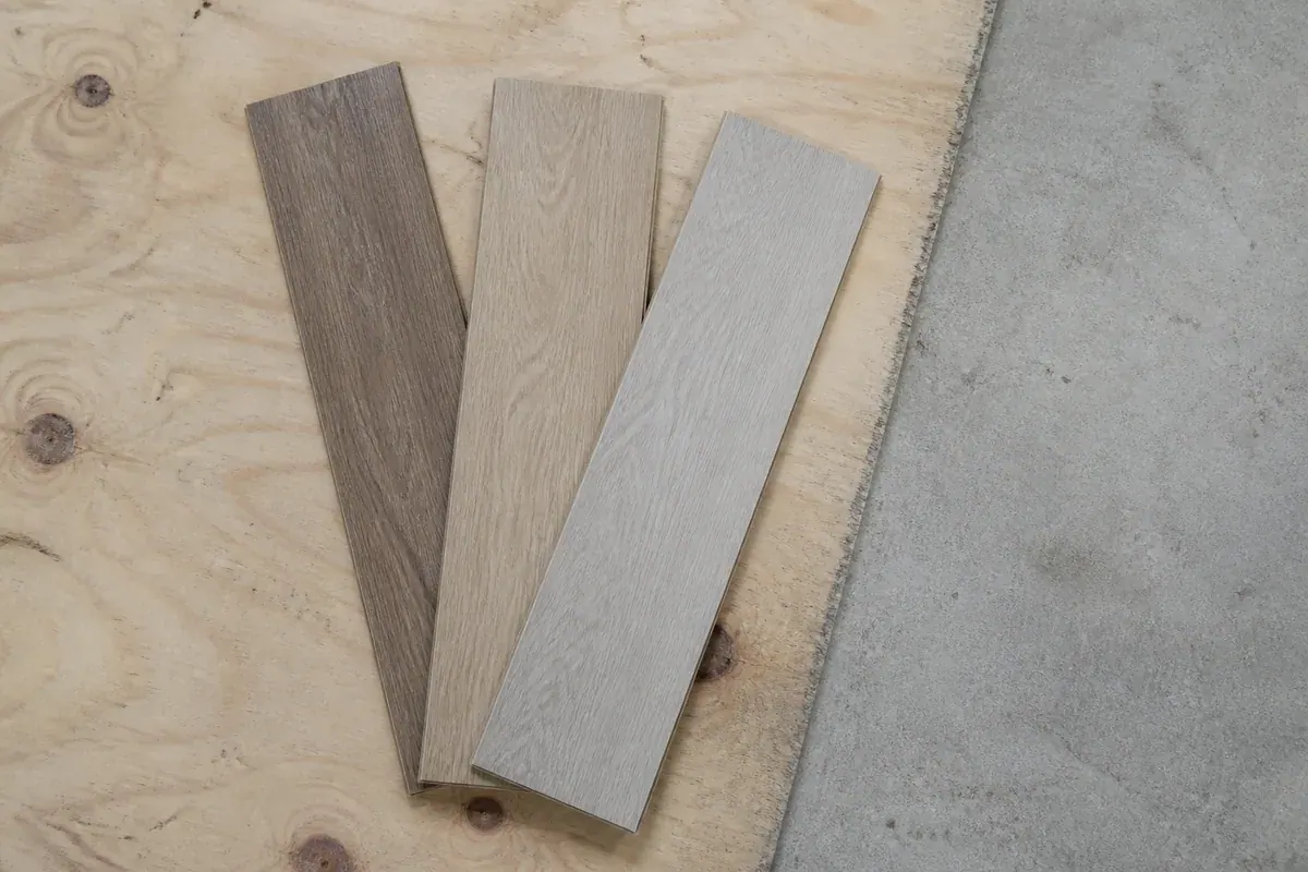Three flooring plank samples laid across an exposed plywood subfloor beside a concrete slab edge