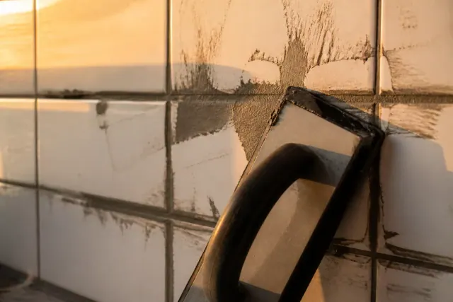 Rubber float pressing gray grout diagonally across the joints of white ceramic wall tile