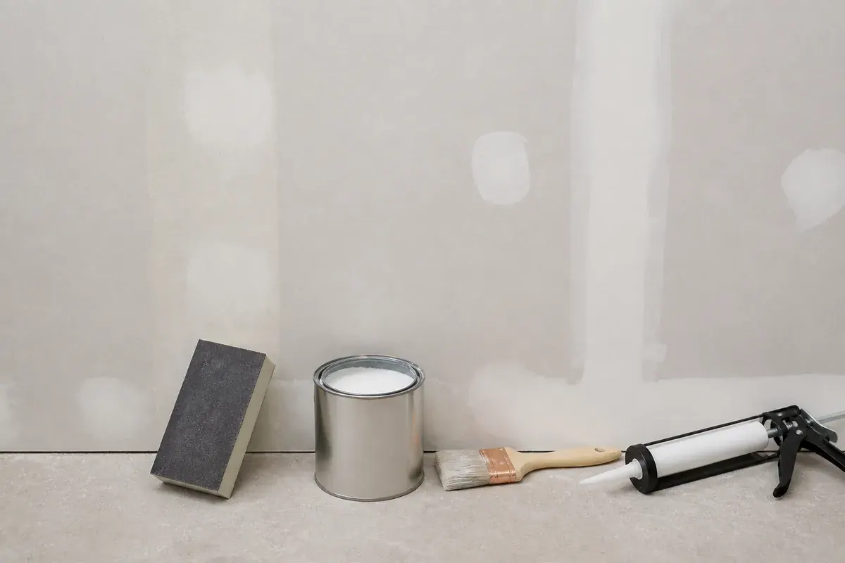 Sanding block smoothing a filled patch on a bare drywall wall next to an open can of white primer, a brush, and a caulk gun