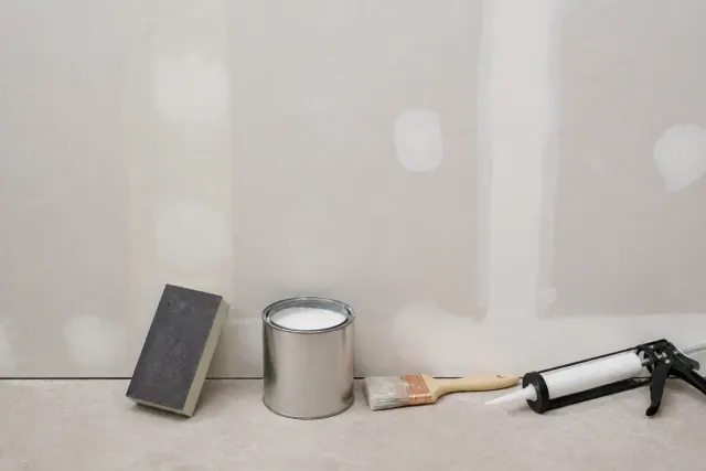 Sanding block smoothing a filled patch on a bare drywall wall next to an open can of white primer, a brush, and a caulk gun