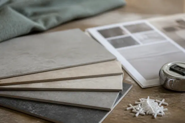 Porcelain floor tile samples fanned across a workbench beside a printed manufacturer specification sheet, a tape measure, and grout spacers